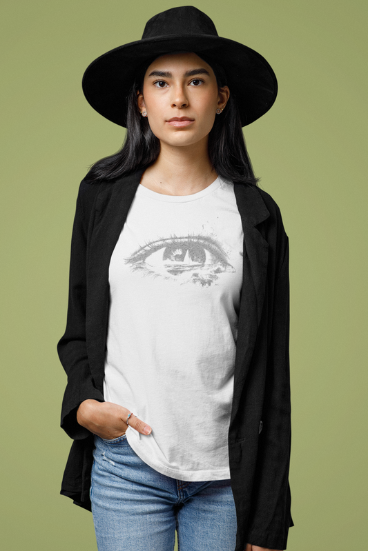 A woman modeling a white graphic t-shirt with a gray eye design, styled with a black blazer, black wide-brim hat, and blue jeans against a solid olive green background.