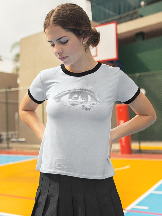 A woman standing on an outdoor basketball court wearing a white ringer T-shirt with black trim and a gray graphic print of an eye, paired with a black pleated skirt.