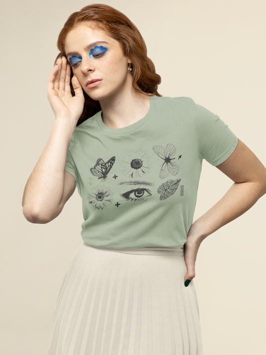A woman wearing a sage green graphic t-shirt with black line drawings of a butterfly, dandelion, dragonfly, and eye, paired with a pleated cream skirt, against a solid beige background.