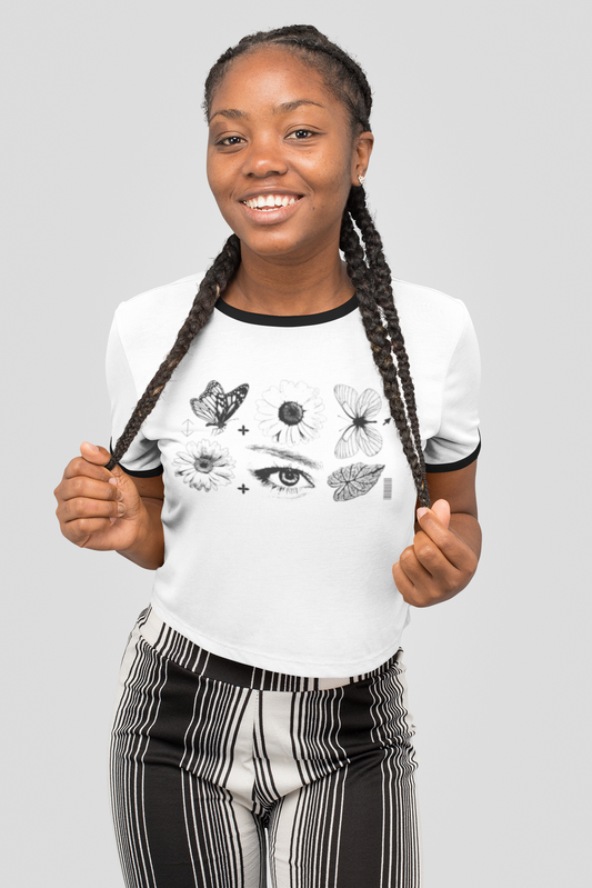 A smiling woman with braided hair wearing a white ringer T-shirt with black graphic prints of butterflies, flowers, and an eye, paired with black and white vertically striped pants, against a solid light gray background.