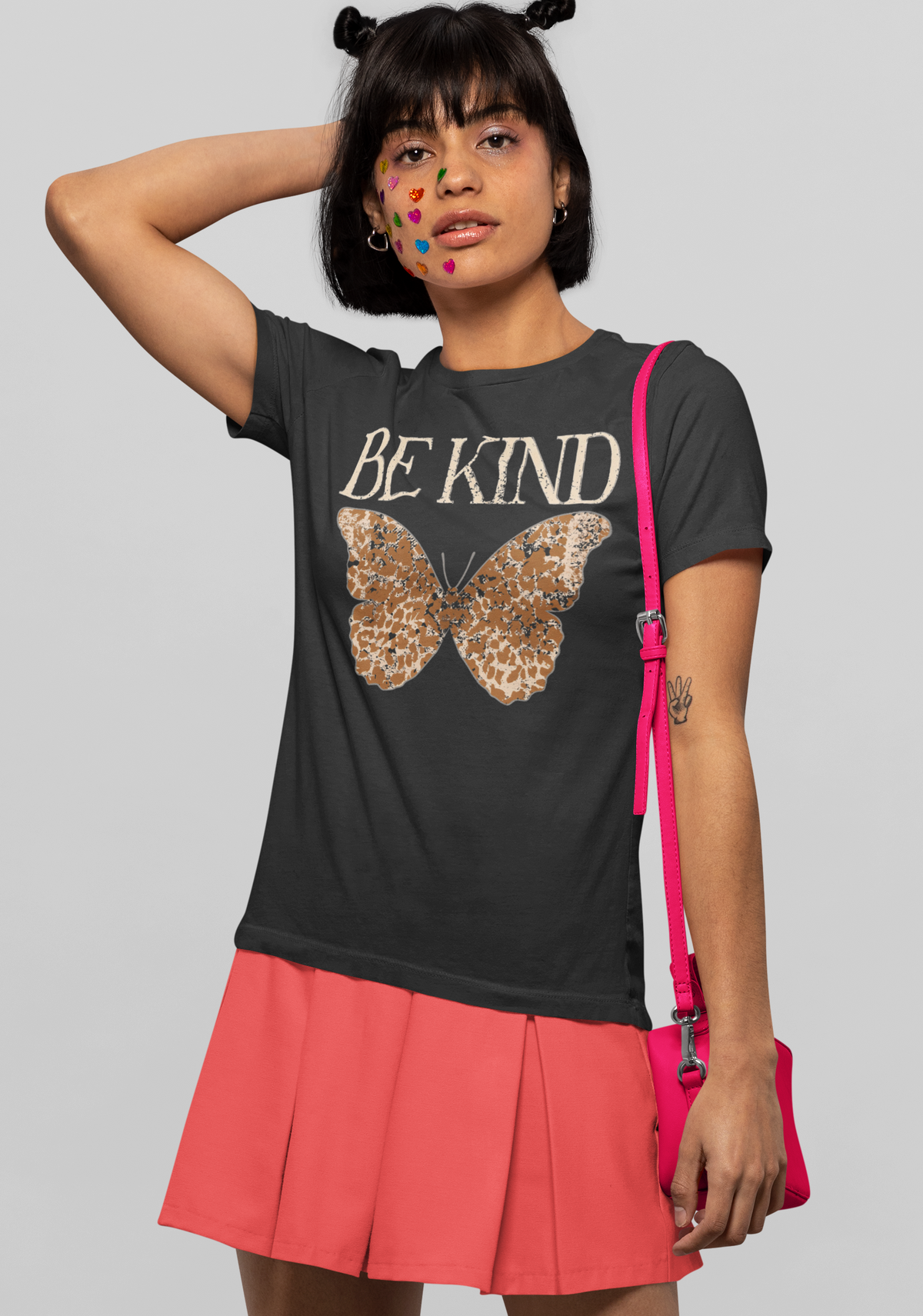 A model wearing a dark gray graphic t-shirt with 'BE KIND' and a butterfly design, paired with a coral pink pleated skirt and a hot pink crossbody bag, against a solid gray background.