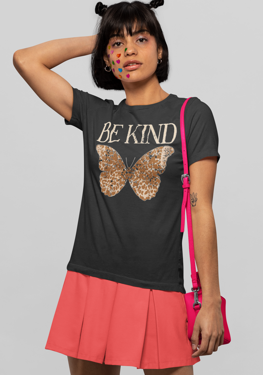 A model wearing a dark gray graphic t-shirt with 'BE KIND' and a butterfly design, paired with a coral pink pleated skirt and a hot pink crossbody bag, against a solid gray background.