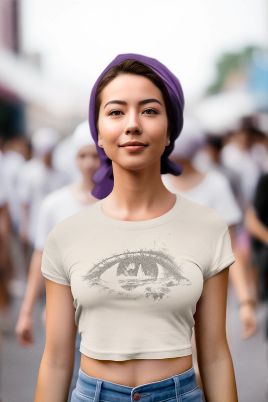 A woman wearing a beige crop top with a detailed eye graphic on the front, paired with blue jeans and a purple headscarf, standing in a blurred outdoor crowd.