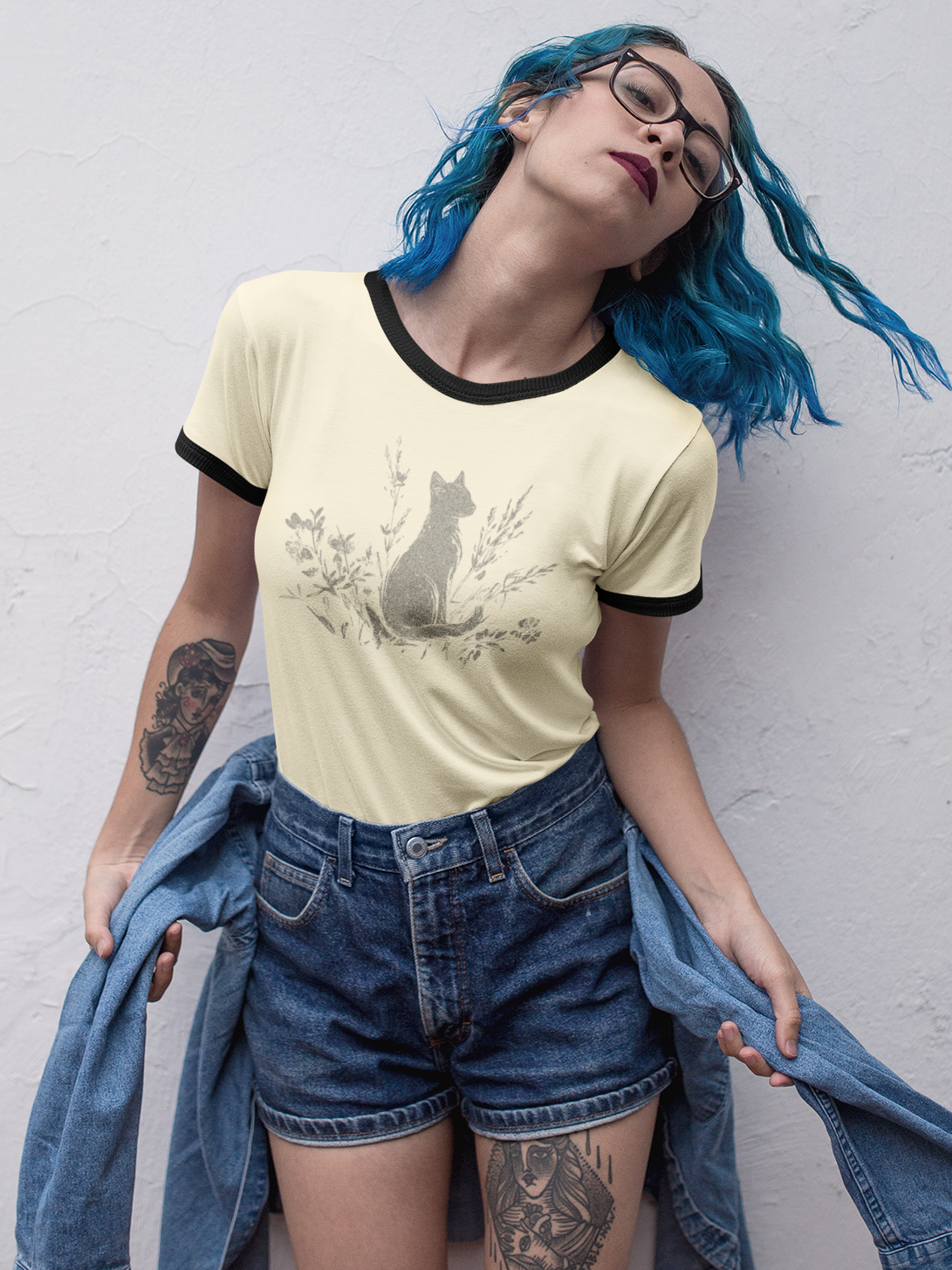 A person with bright blue hair and glasses poses against a white textured wall, wearing a pale yellow ringer T-shirt with a black collar and sleeve trim, a gray cat graphic, blue denim shorts, and holding a blue denim jacket.