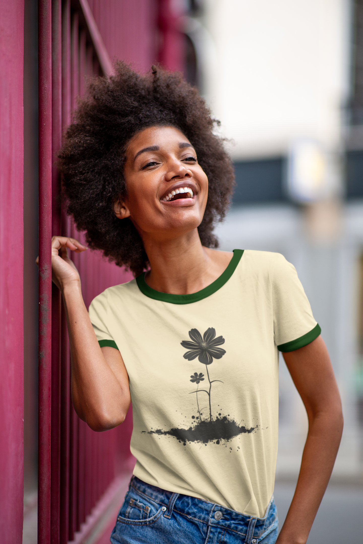 A woman with an afro hairstyle smiling and looking upward while wearing a cream ringer t-shirt with green trim and a black flower graphic, paired with blue jeans, standing next to a dark magenta metal structure.